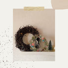 Auf einem dekorativen Regal stehen ein Zweigkranz, ein herzförmiges Ornament mit pastellfarbener Stanzschablone Rosenblüte von Stanzenshop.de und ein kleiner grüner Flaschenbürstenbaum - perfekt, um jeder schlichten Wand Scrapbooking-Charme zu verleihen.