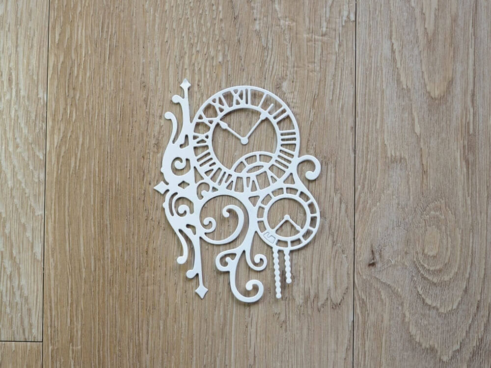 Weiße dekorative Uhrenzahnräder und Schnörkel, die mit der Stanzschablone Zwei Uhren mit Ranken von Stanzenshop.de gefertigt wurden, werden auf einer hellbraunen Holzoberfläche dargestellt und bilden ein inspirierendes Bastelergebnis, das sich ideal für Scrapbooking-Projekte eignet.