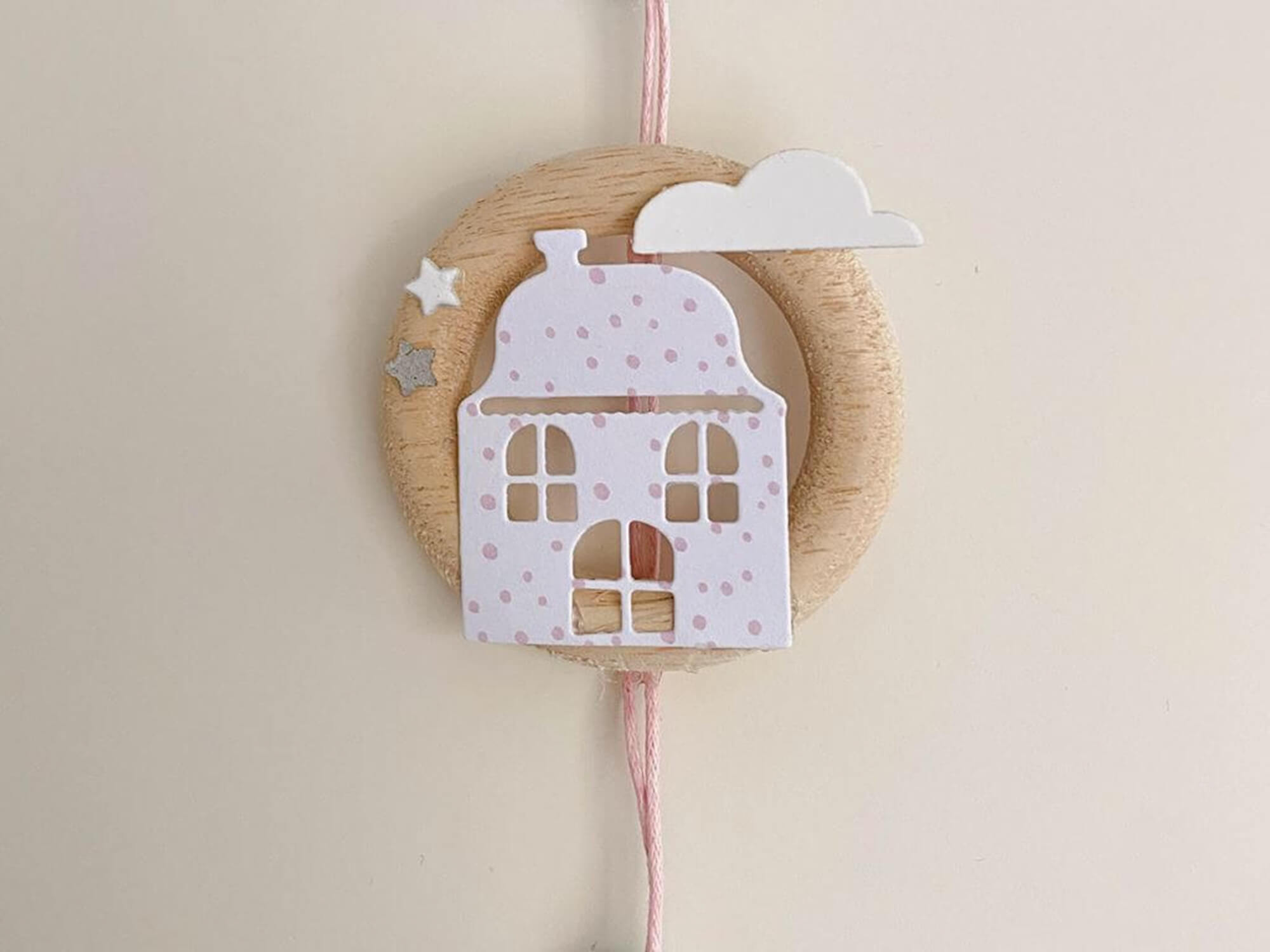 Ein rundes Holzornament mit rosa gepunktetem Haus, weißer Wolke und Sternen hängt an einer rosa Schnur - perfekt, um die "Stanzschablone: Zwei verschiedene Häuser" von Stanzenshop.de als Highlight vor hellen Hintergründen.