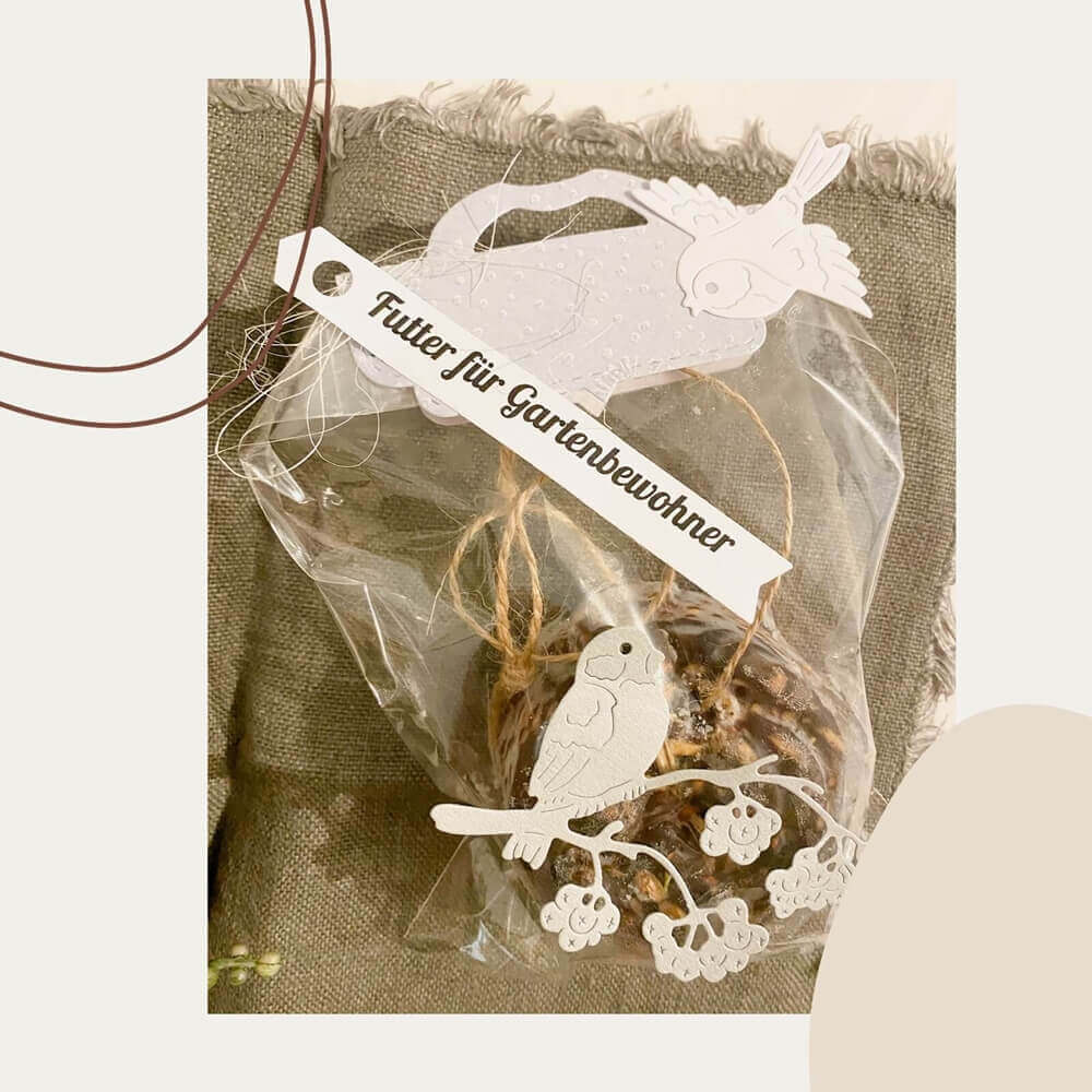 Eine Verpackung mit Vogelfutter mit dekorativen Papierausschnitten von Vögeln, hergestellt mit der Stanzschablone Verschluss für Tüten von Stanzenshop.de, und einem angebrachten Etikett mit der Aufschrift „futter für gartenbewohner“.