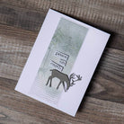 Eine handgefertigte Karte mit einer silbernen Rentier-Silhouette, die mit den Sizzix 664968 Thinlits Darling Deer von Tim Holtz ausgestanzt wurde, dem Text "Friede auf Erden" und einem strukturierten Hintergrund - ideal für die Kartenherstellung. Ausgestellt auf Holz. Marke: Sizzix.