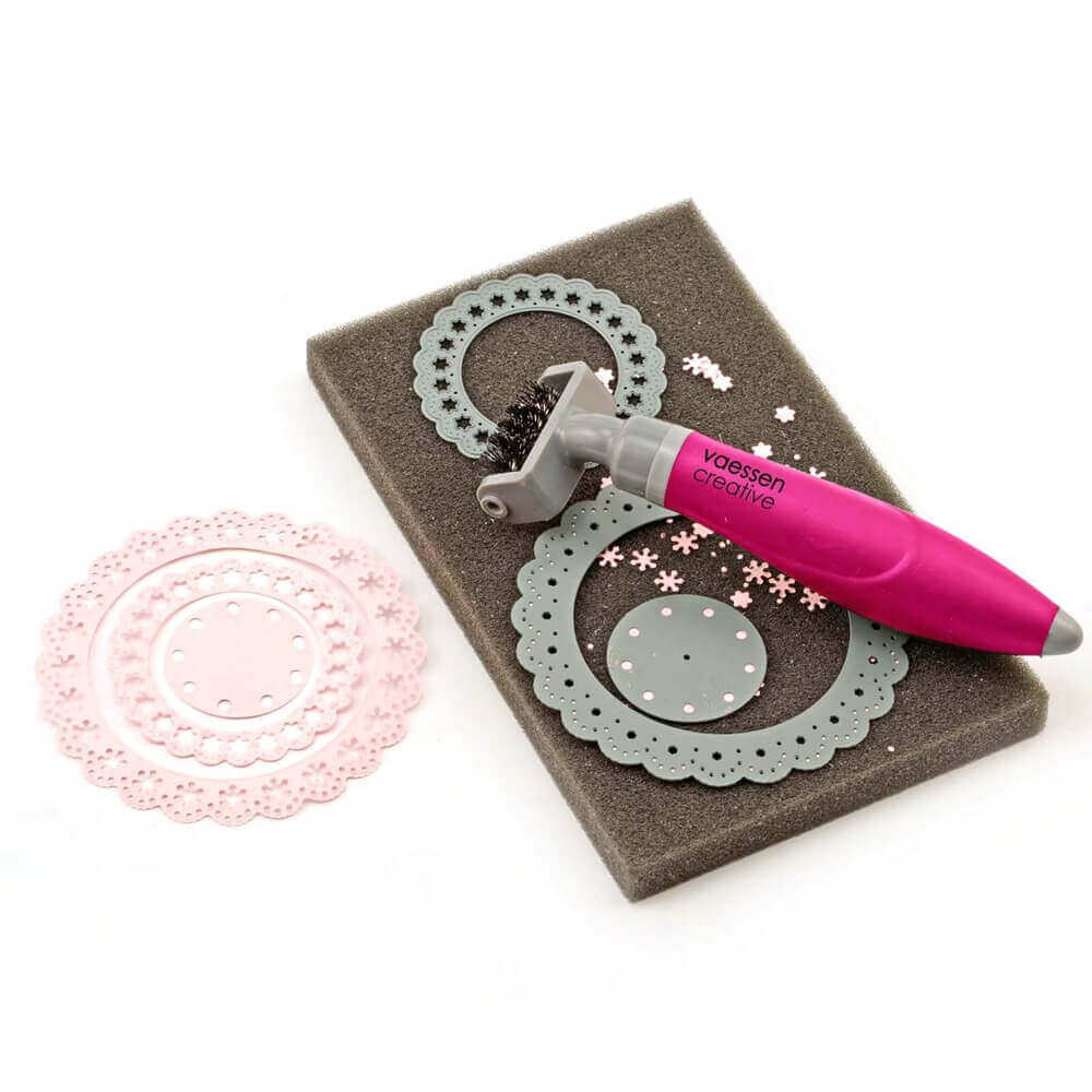 Die Vaessen Creative Die brush & foam pad von Vaessen Creative hat einen rosafarbenen Griff und wird verwendet, um bei der Reinigung von Metallstanzen kleine Papierstücke von komplizierten runden Stanzmustern zu entfernen.