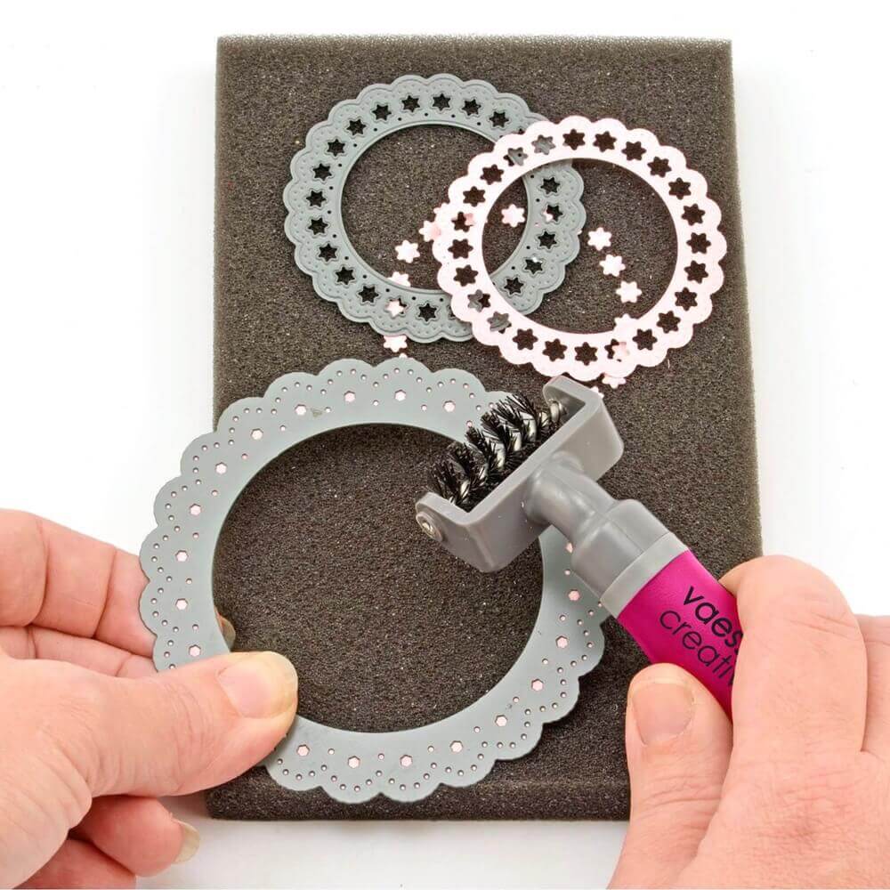 Mit dem Vaessen Creative Die Pinsel und dem Schaumstoffkissen entfernt eine Person kleine Papierpunkte von einem runden dekorativen Metallstempel, der auf dem Schaumstoffkissen platziert ist, wobei zwei ähnliche Stempel in der Nähe liegen.