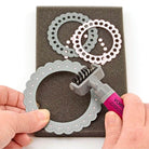 Mit dem Vaessen Creative Die Pinsel und dem Schaumstoffkissen entfernt eine Person kleine Papierpunkte von einem runden dekorativen Metallstempel, der auf dem Schaumstoffkissen platziert ist, wobei zwei ähnliche Stempel in der Nähe liegen.