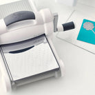 Eine Sizzix Big Shot mit der Sizzix 660470 Chrome Precision Base Plate für komplizierte Thinlits liegt auf einem Tisch neben einer blauen Karte mit einem weißen Blumenausschnitt und demonstriert die perfekte Schnittqualität.