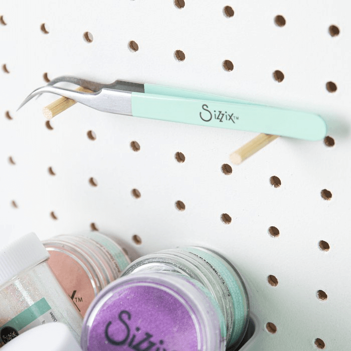 Die Sizzix 664140 Werkzeug gebogene Pinzette mit feiner Spitze von Sizzix hängt an einem von zwei Dübeln gehaltenen Steckbrett, unter dem sich Glitzerdosen befinden. Ihre Produktabmessungen und Verpackungsmaße sorgen dafür, dass sie perfekt in Ihren Bastelbereich passt.